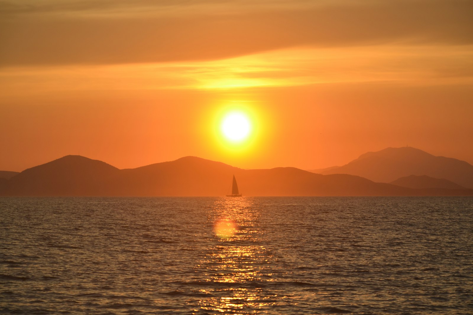 A sailboat on the Aegean at sunset with the sun low over island hills